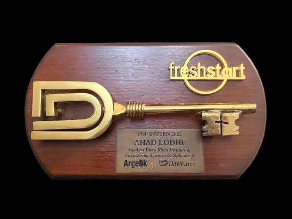 A brass key mounted on a wooden plaque beside the Dawlance D monogram and a freshstart wordmark. The plate beneath reads Top Intern 2022, Ahad Lodhi, Ghulam Ishaq Khan Institute of Engineering Sciences and Technology, with the Arcelik and Dawlance marks.