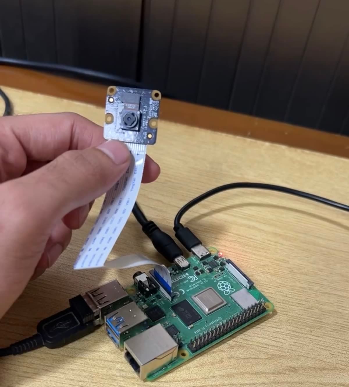 A hand holding a Raspberry Pi camera module by its ribbon cable above a Raspberry Pi 4 board wired up on a desk.